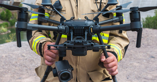 A firefighter holding a black drone by the landing gear standing in front of a tall, concrete dam.